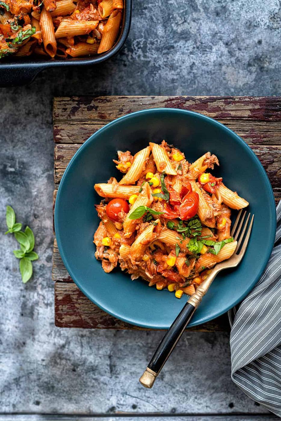 A bowl of penne pasta with tomato sauce, cherry tomatoes, corn, and herbs reminds one of a deconstructed tuna pasta bake. Placed on a wooden surface, a fork rests beside it while fresh basil leaves add a burst of color. A striped cloth is partially visible on the side.