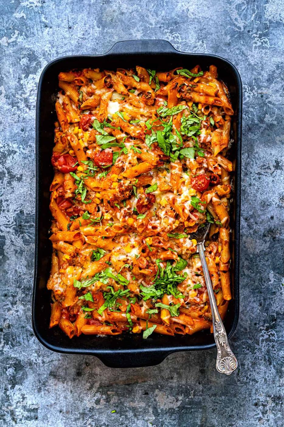 A black baking dish filled with cheesy baked penne pasta, topped with a rich tomato sauce, fresh basil, and sprinkled with melted cheese. A metal serving spoon rests in the dish, all set on a textured gray surface.