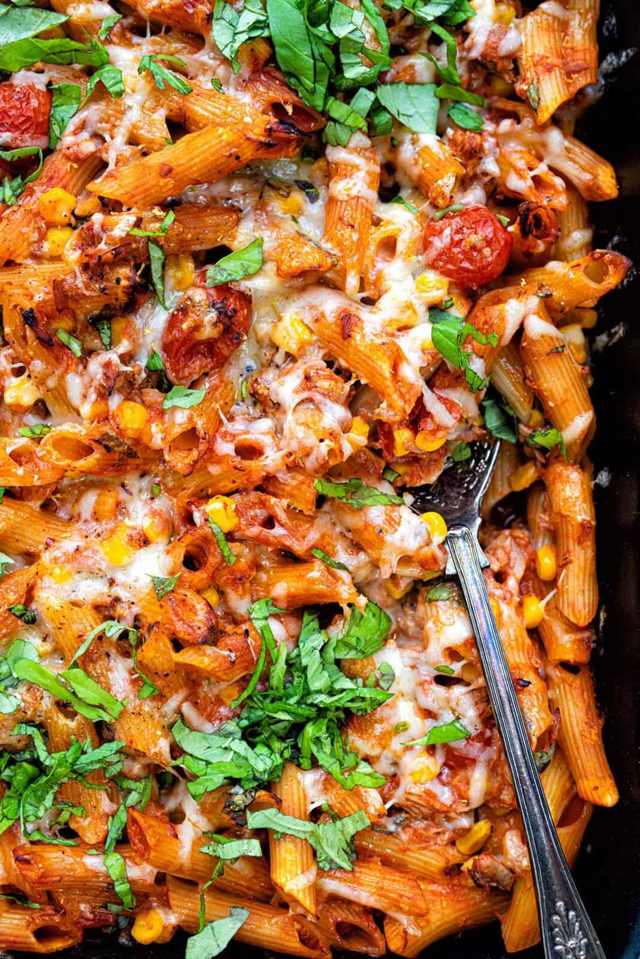 A close-up of a healthy tuna casserole showcasing penne, cherry tomatoes, corn, melted cheese, and garnished with fresh basil. A serving spoon is partially submerged in the cheesy pasta.