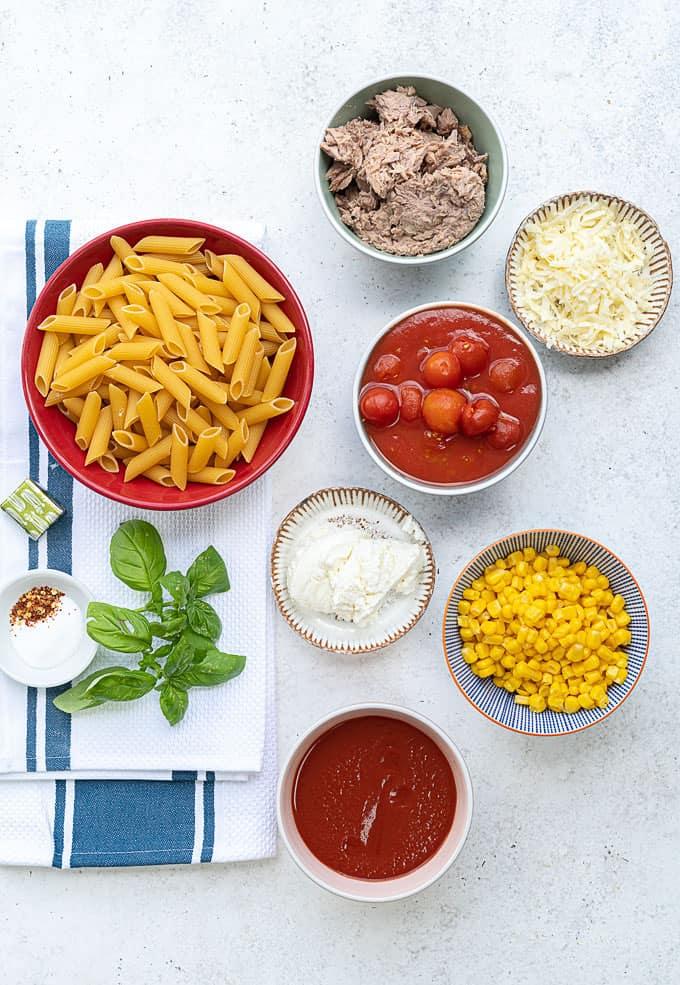 A flat lay of pasta ingredients for a slimming world pasta bake: dry penne, shredded beef, shredded cheese, ricotta, cherry tomatoes in sauce, corn, tomato sauce, basil leaves, a stock cube, salt, and chili flakes on a white surface with a blue-striped towel.