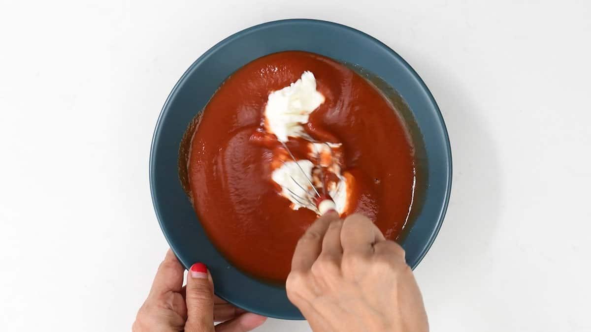 A person whisking a dollop of cream or yogurt into a bowl of red tomato soup, much like preparing a creamy base for a healthy tuna casserole, with both hands holding the bowl and whisk.
