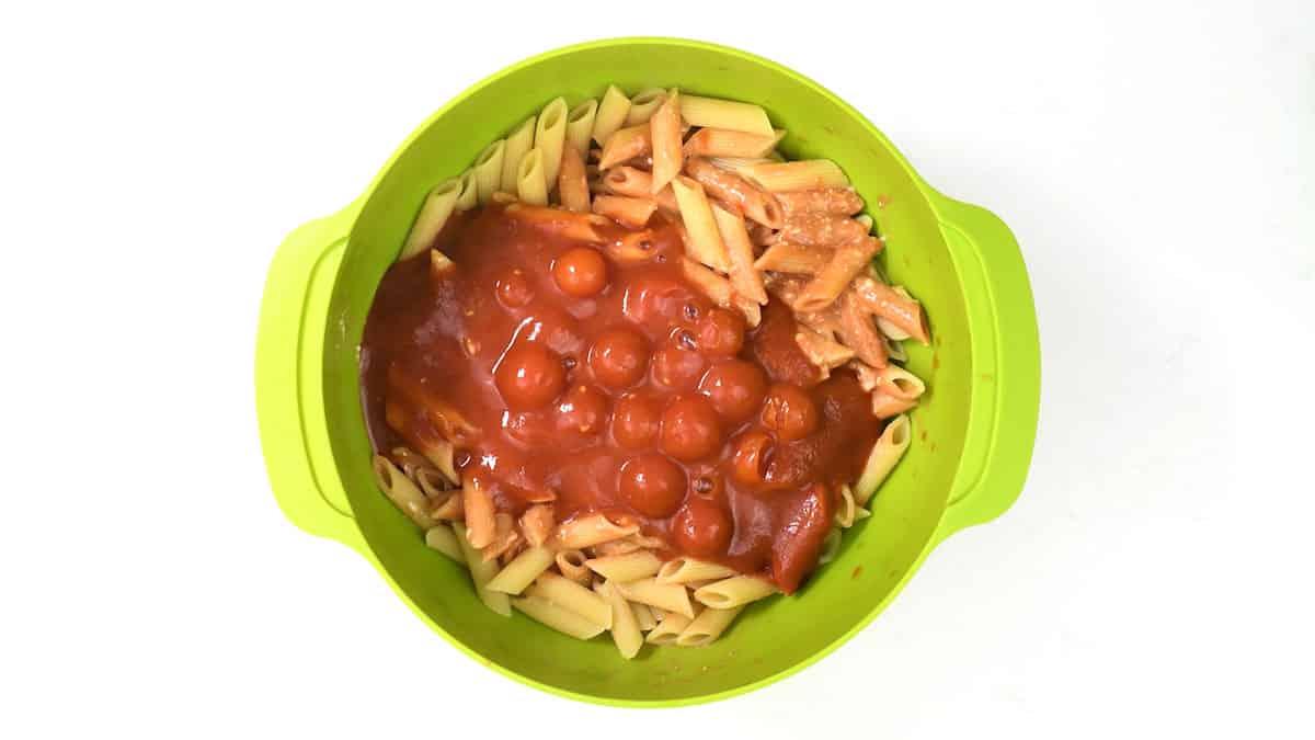 A green bowl brimming with cooked penne pasta is topped with a chunky red tomato sauce. The sauce, enriched with cherry tomatoes, adds vibrant contrast. With handles on either side, this dish resembles a classic tuna pasta bakes hearty appeal.
