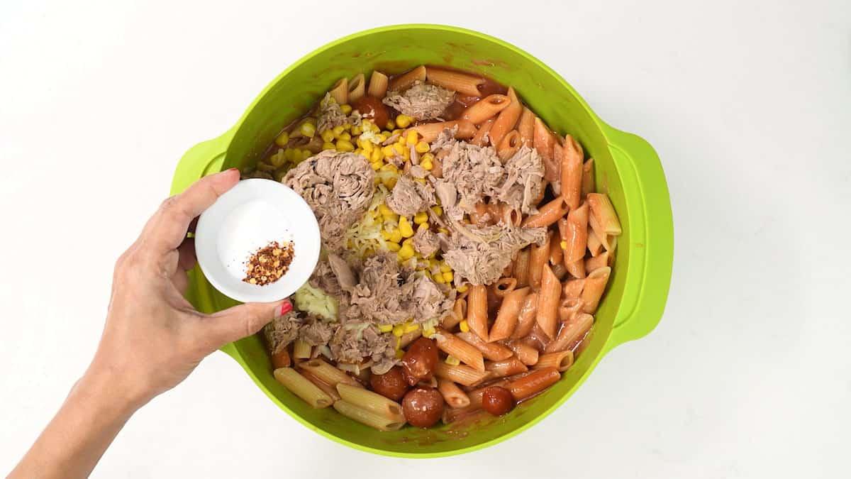 A person is holding a small white bowl of red pepper flakes over a large green bowl filled with pasta, tuna, corn, and cherry tomatoes. These ingredients are being prepared for mixing into a delicious tuna pasta bake.