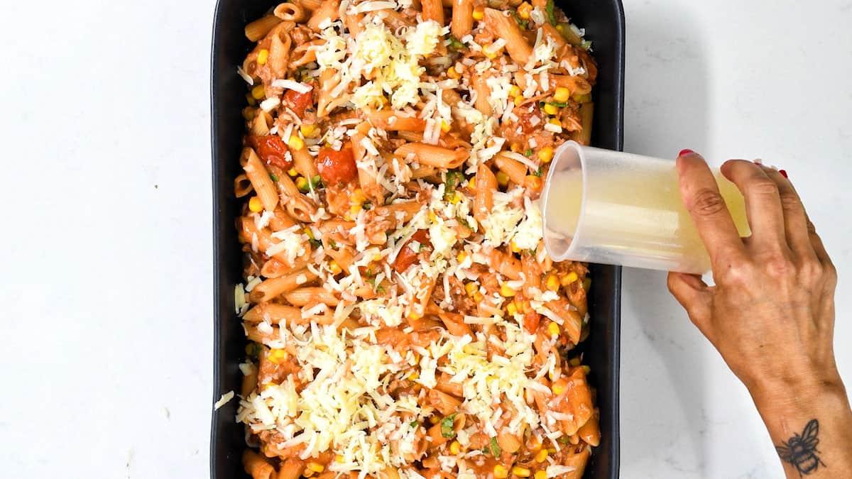 A hand pours a creamy liquid over a healthy tuna casserole dish filled with penne pasta, vegetables, and shredded cheese on a white surface.