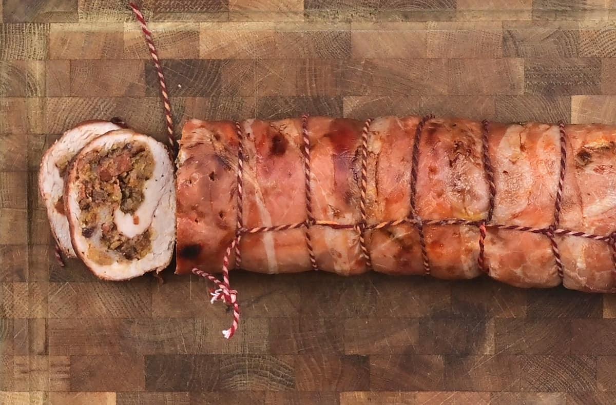 A boned and rolled turkey breast with stuffing, wrapped in prosciutto, tied with twine and sliced to reveal a savory filling, sits on a wooden cutting board.