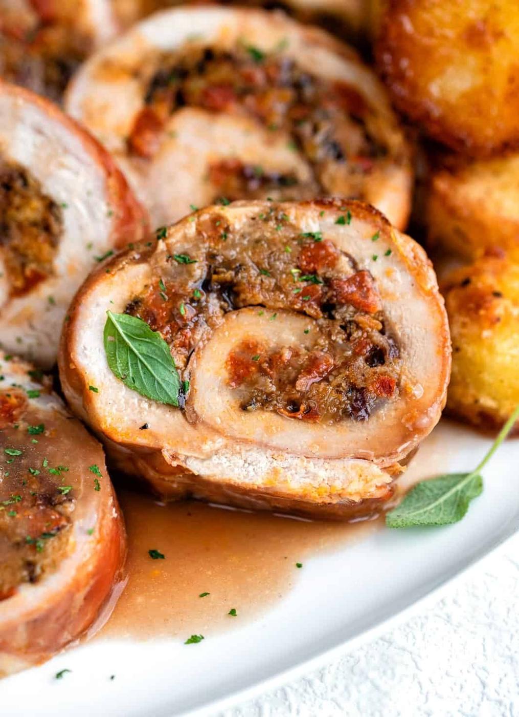 Close-up of sliced turkey roll roulade with a savory filling, garnished with fresh herbs and served alongside golden roasted potatoes on a white plate.
