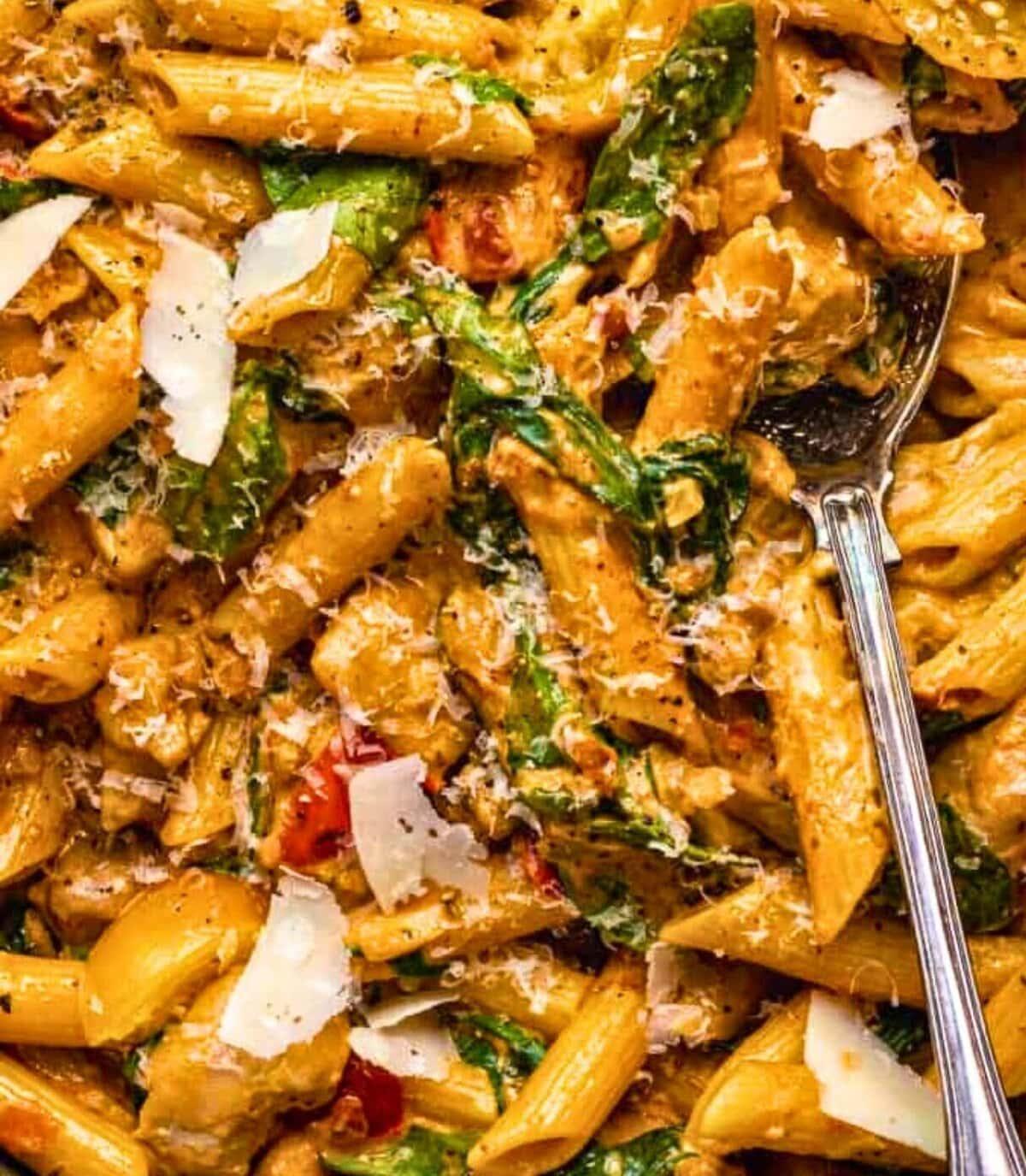 Close-up of creamy Tuscan Chicken Pasta with spinach, sun-dried tomatoes, and shaved parmesan, with a silver spoon. Above, elegant text reads “Festive Cocktail THE RED QUEEN.”.