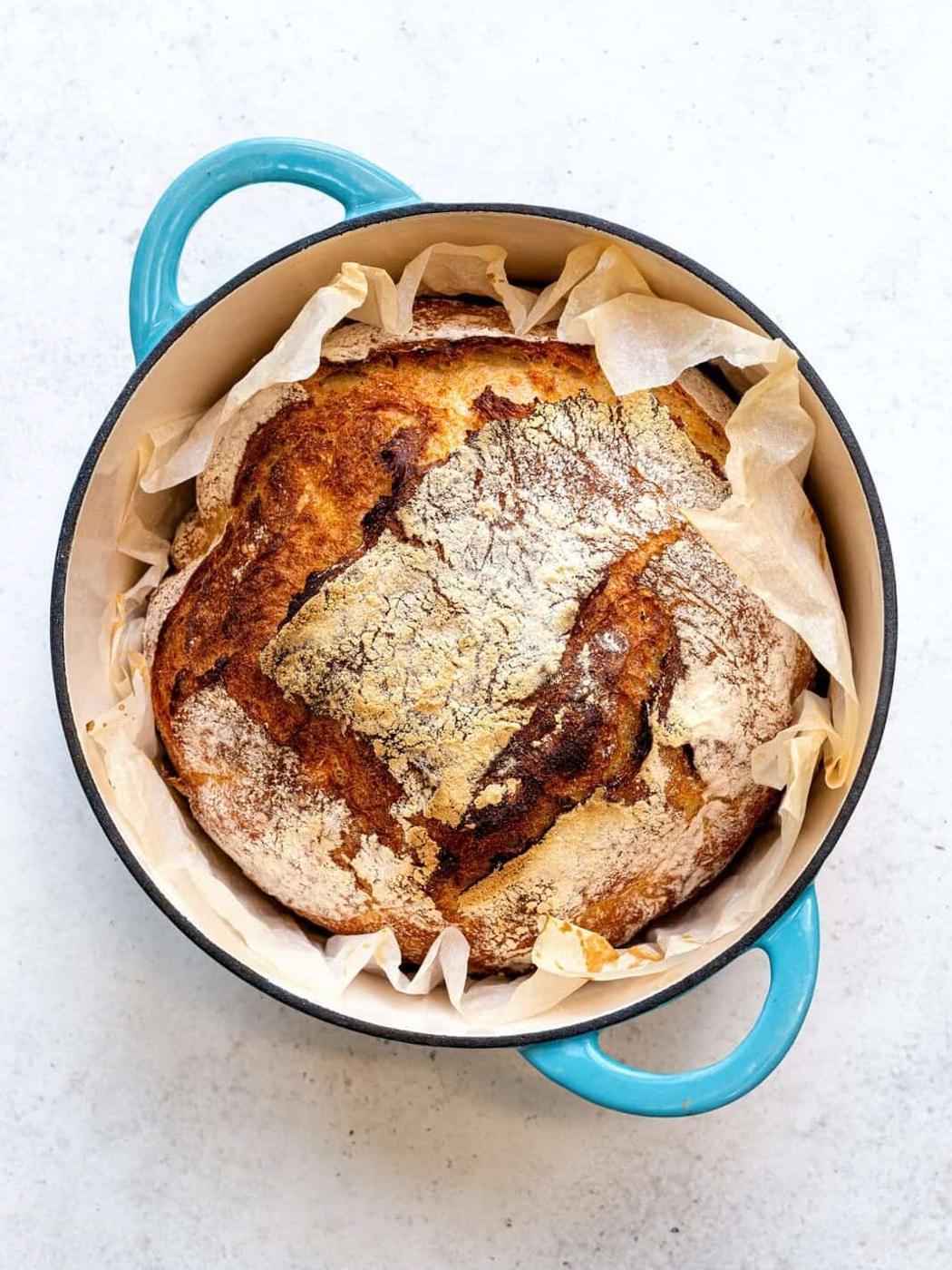 A freshly baked loaf of no-knead bread sits in a blue Dutch oven lined with parchment paper, showcasing a golden-brown crust and rustic texture. The background is a light, neutral surface.