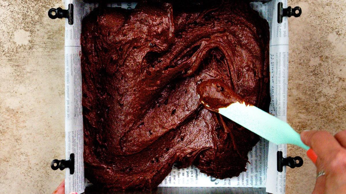 A hand uses a light blue spatula to spread thick chocolate brownie batter in a square baking pan lined with parchment paper printed with text.