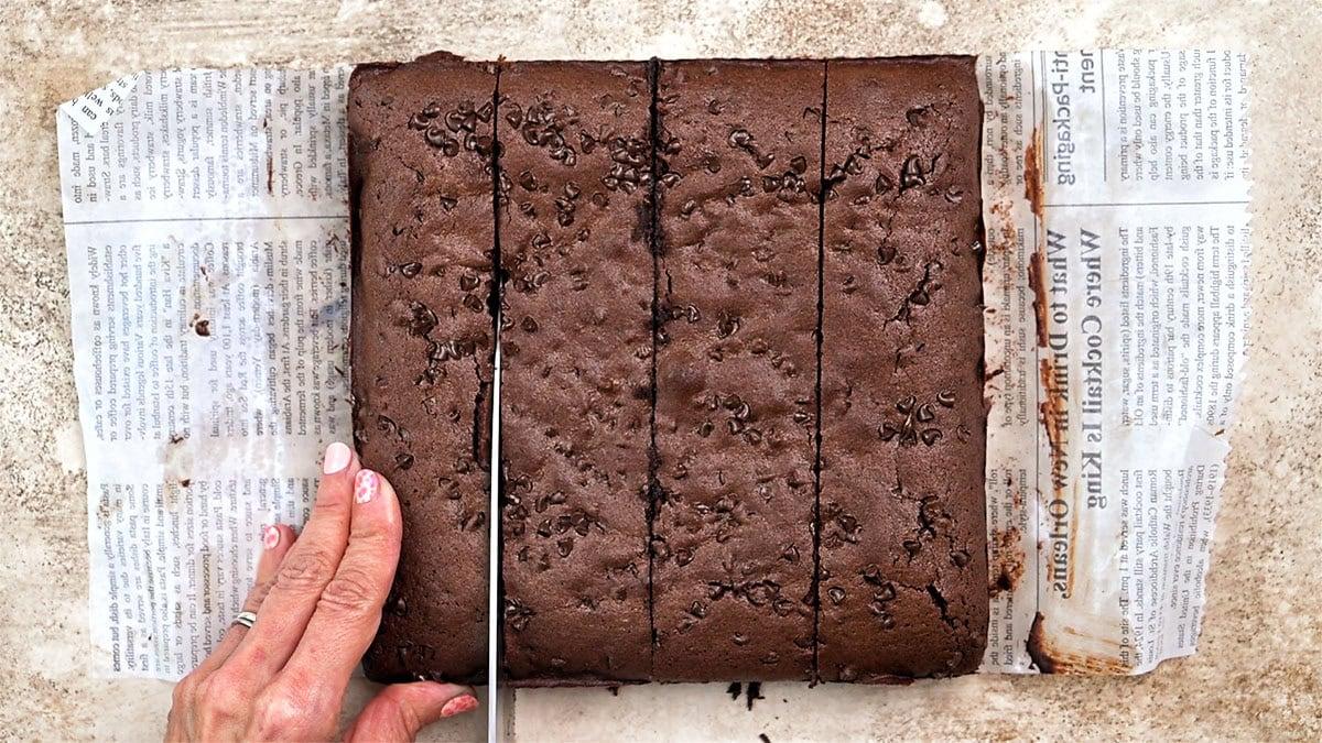 A hand is cutting a large, square chocolate brownie topped with chocolate chips into pieces on a sheet of newspaper, placed on a light-colored textured surface.