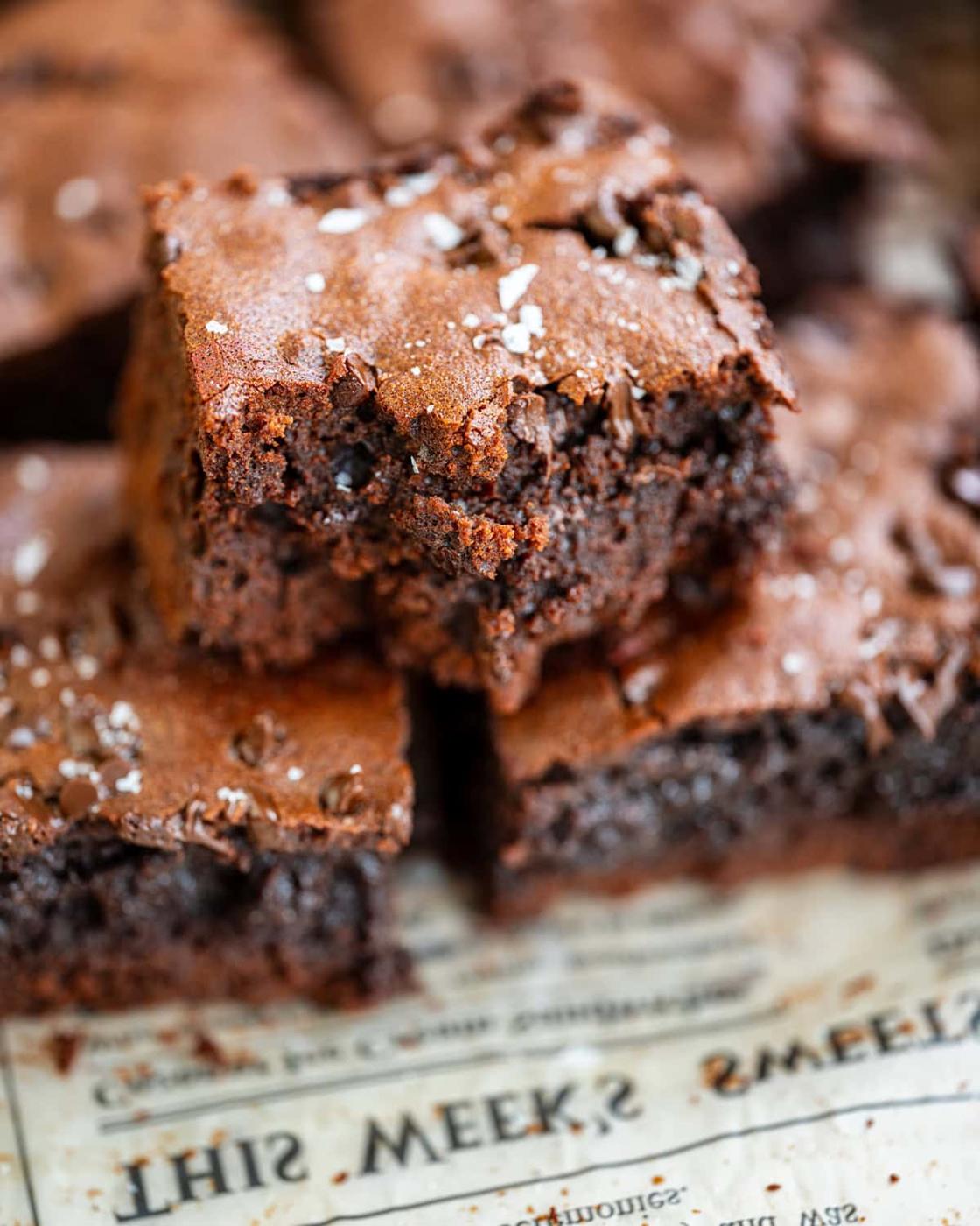 A close-up of chocolate protein brownies stacked on old newspaper, with one brownie showing a bite taken out. The brownies have a rich, fudgy texture and are sprinkled with flaky sea salt.