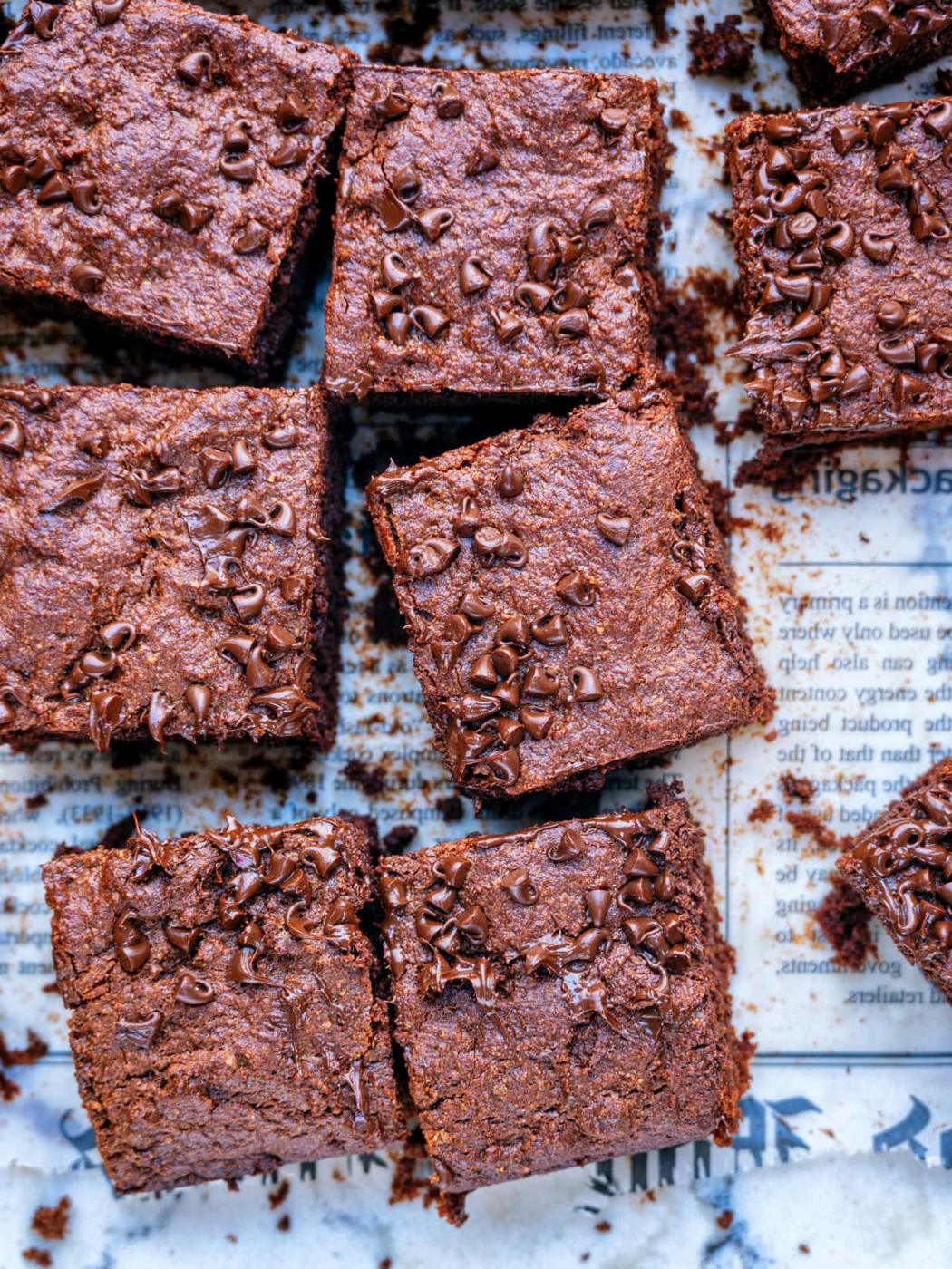 Nine chocolate protein brownies topped with mini chocolate chips are arranged on a piece of parchment paper printed with text. The brownies appear moist and fudgy, with crumbs scattered around.