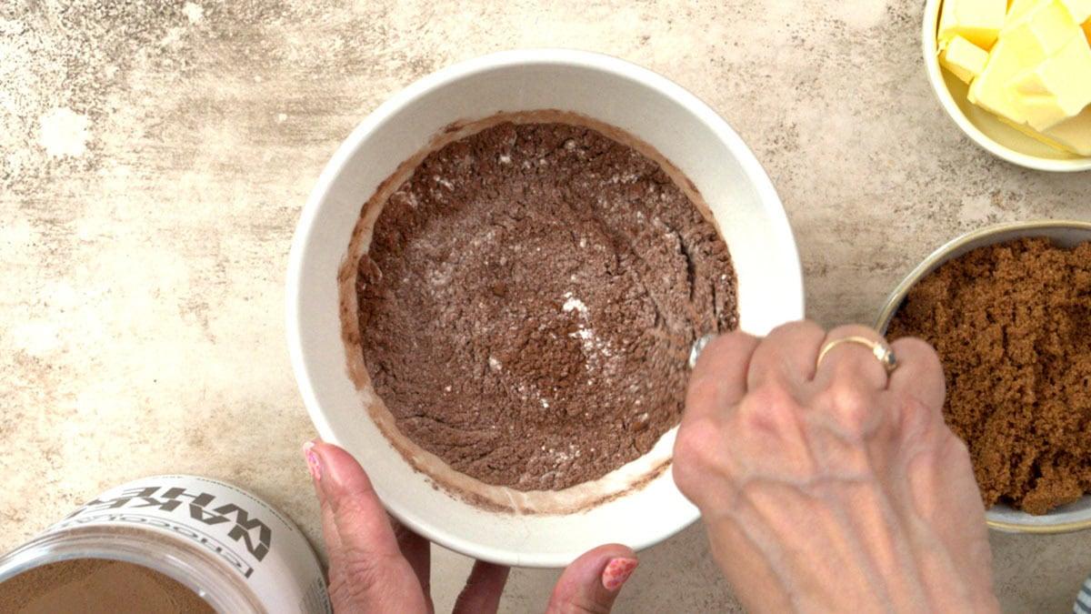 A person stirs a bowl of dry cocoa powder mixture on a countertop. Nearby are bowls containing brown sugar and butter, and a container labeled Naked Nut.
