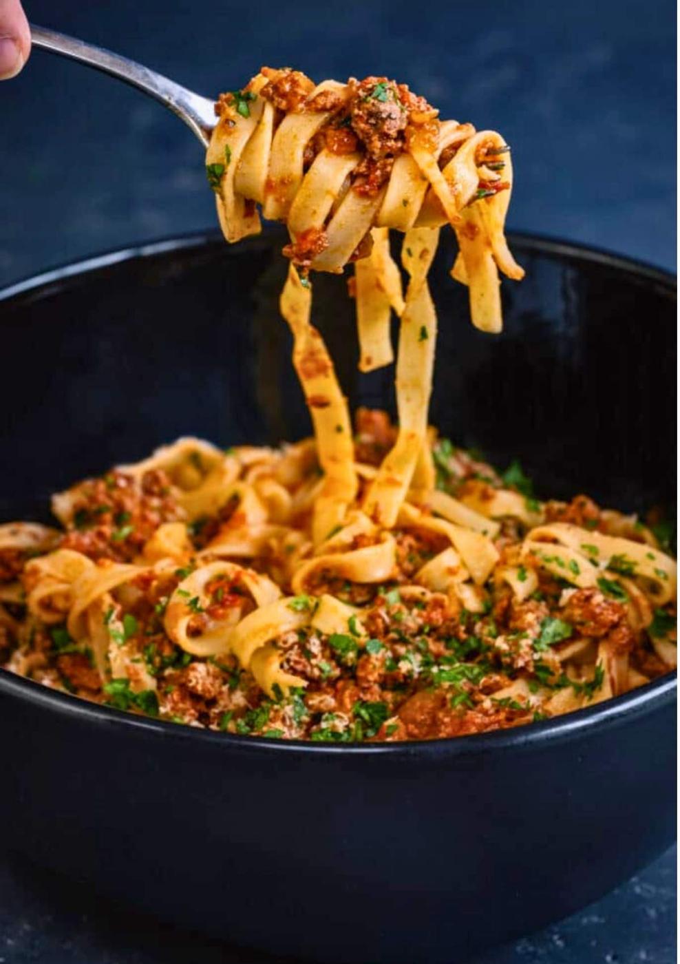 A fork lifts a portion of tagliatelle pasta topped with slow cooked venison ragu from a black bowl filled with more pasta and sauce, garnished with chopped herbs.