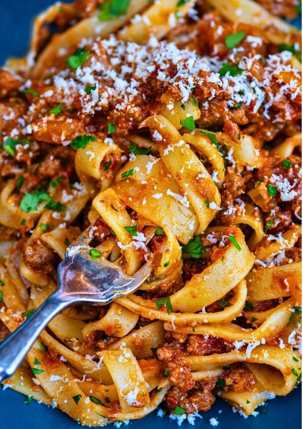 A close-up of tagliatelle pasta with slow cooked ground venison ragu, topped with grated Parmesan cheese and chopped parsley, with a fork twirling some noodles.