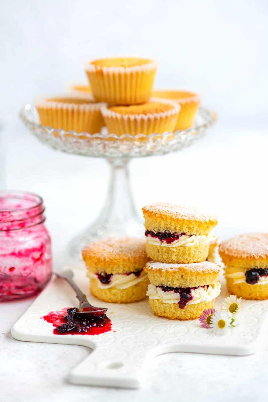 Mini Victoria sponge cupcakes filled with cream and berry jam are stacked on a white board, with a jar of jam, a knife, and a glass stand holding more cupcakes in the background. Small flowers decorate the scene.