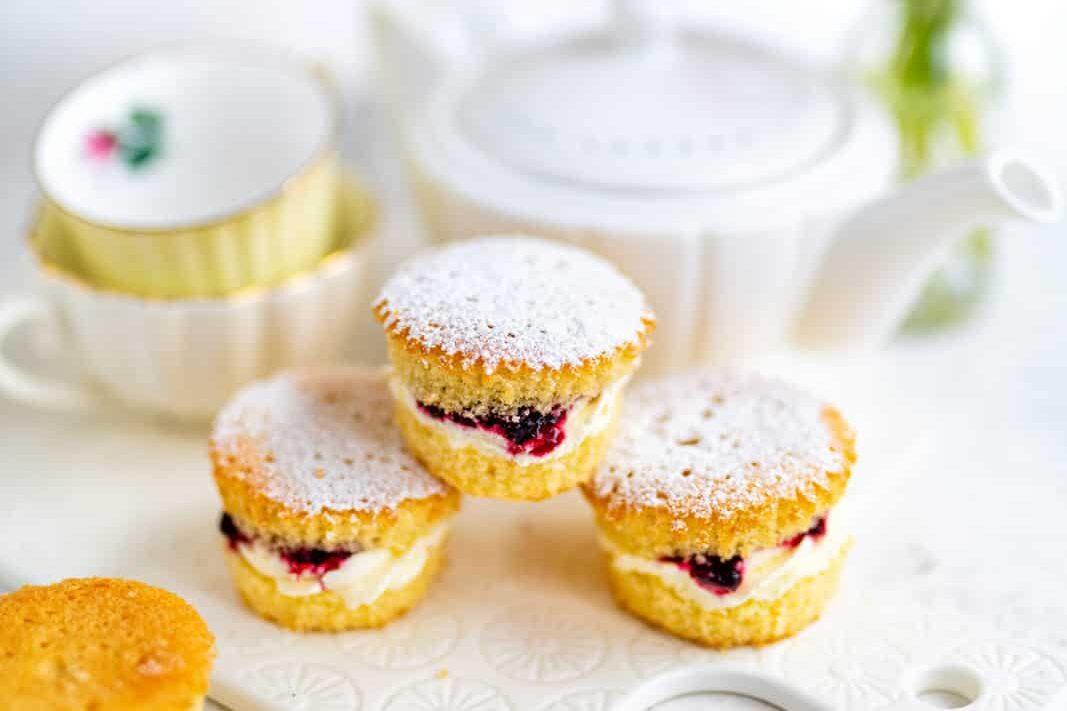 Three small Victoria sponge cakes filled with cream and jam are dusted with powdered sugar. They are arranged on a white plate with a teapot and stacked teacups in the blurred background.