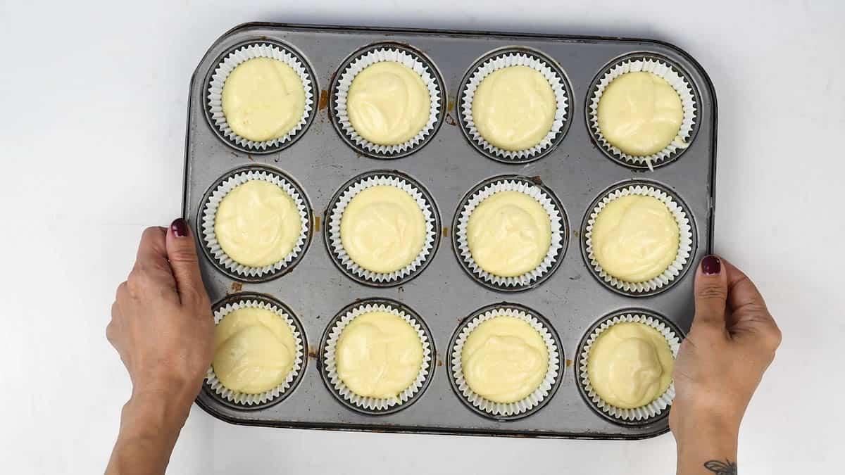A person holds a muffin tray filled with white cupcake liners, each containing unbaked Victoria sponge cupcakes batter, against a white background.
