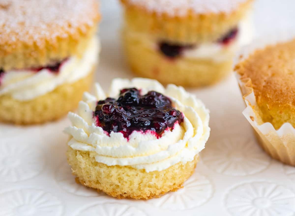 A close-up of Victoria sponge cupcakes, one topped with whipped cream and berry jam, others filled with cream and jam, on a patterned white surface.