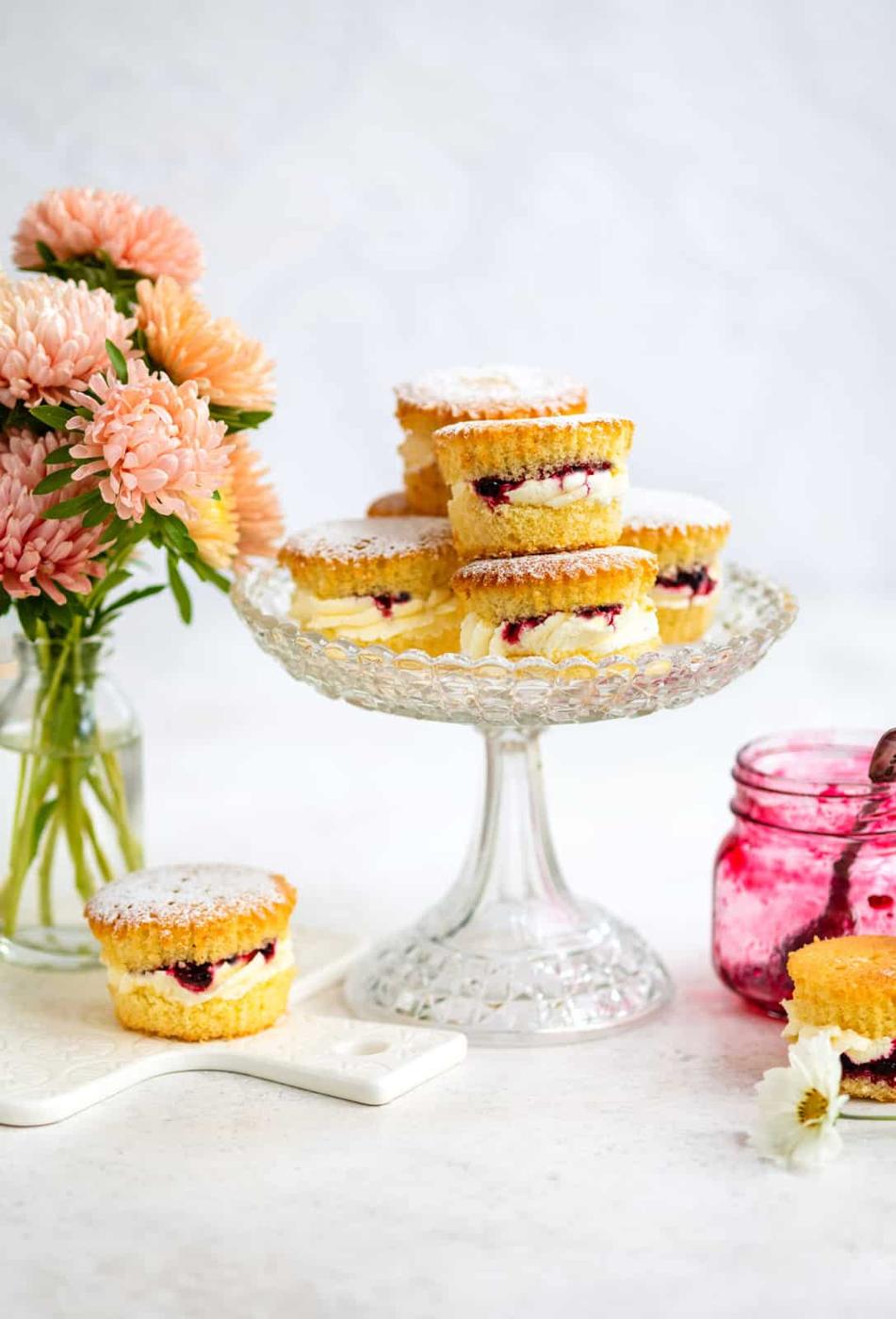 Mini Victoria sponge cupcakes filled with cream and jam are stacked on a glass cake stand. A jar of jam, a vase of pink flowers, and a single mini cake are arranged on the white surface nearby.