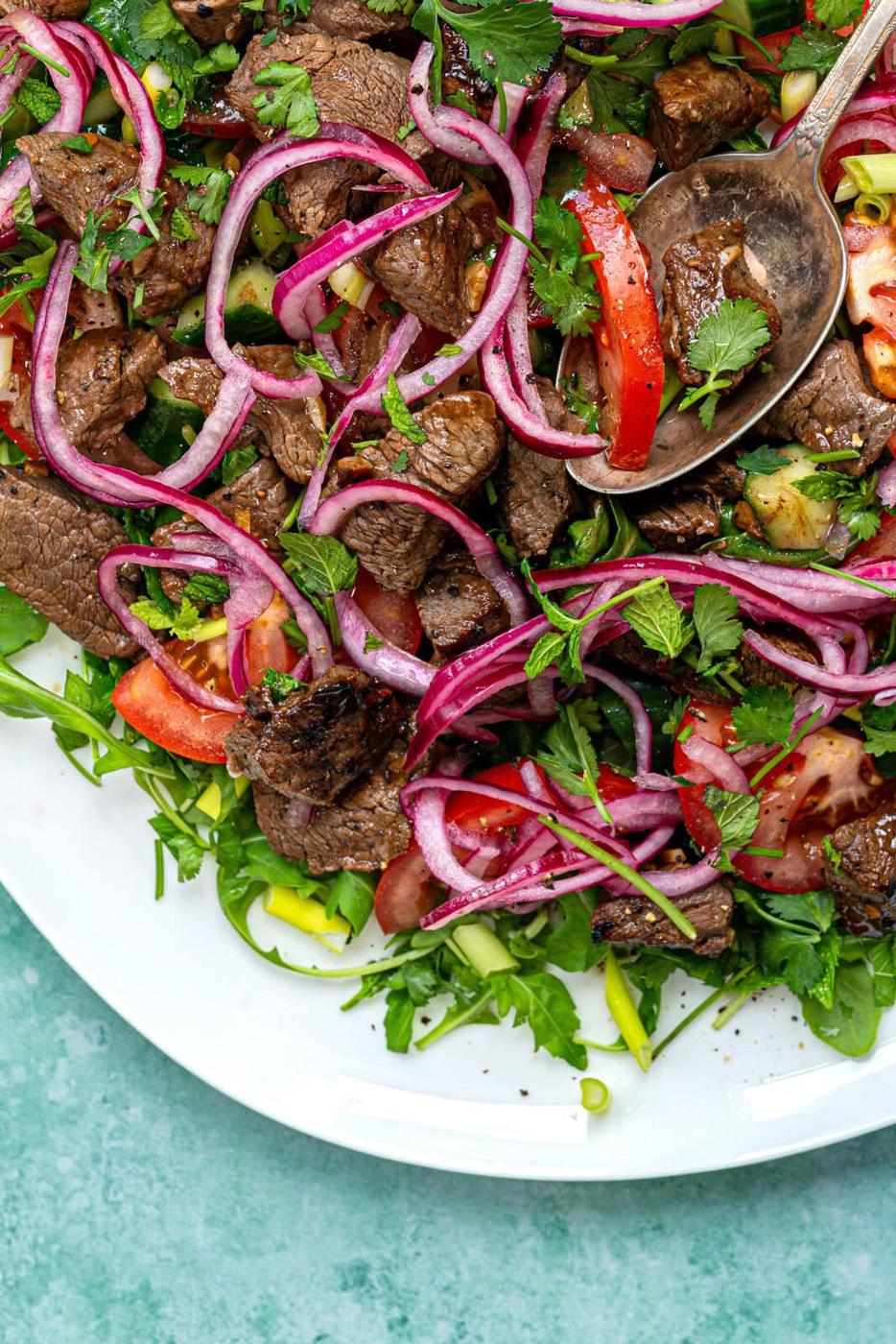 A close-up of a salad with Vietnamese shaking, red onion slices, tomatoes, fresh herbs, and greens on a white plate with a serving spoon.