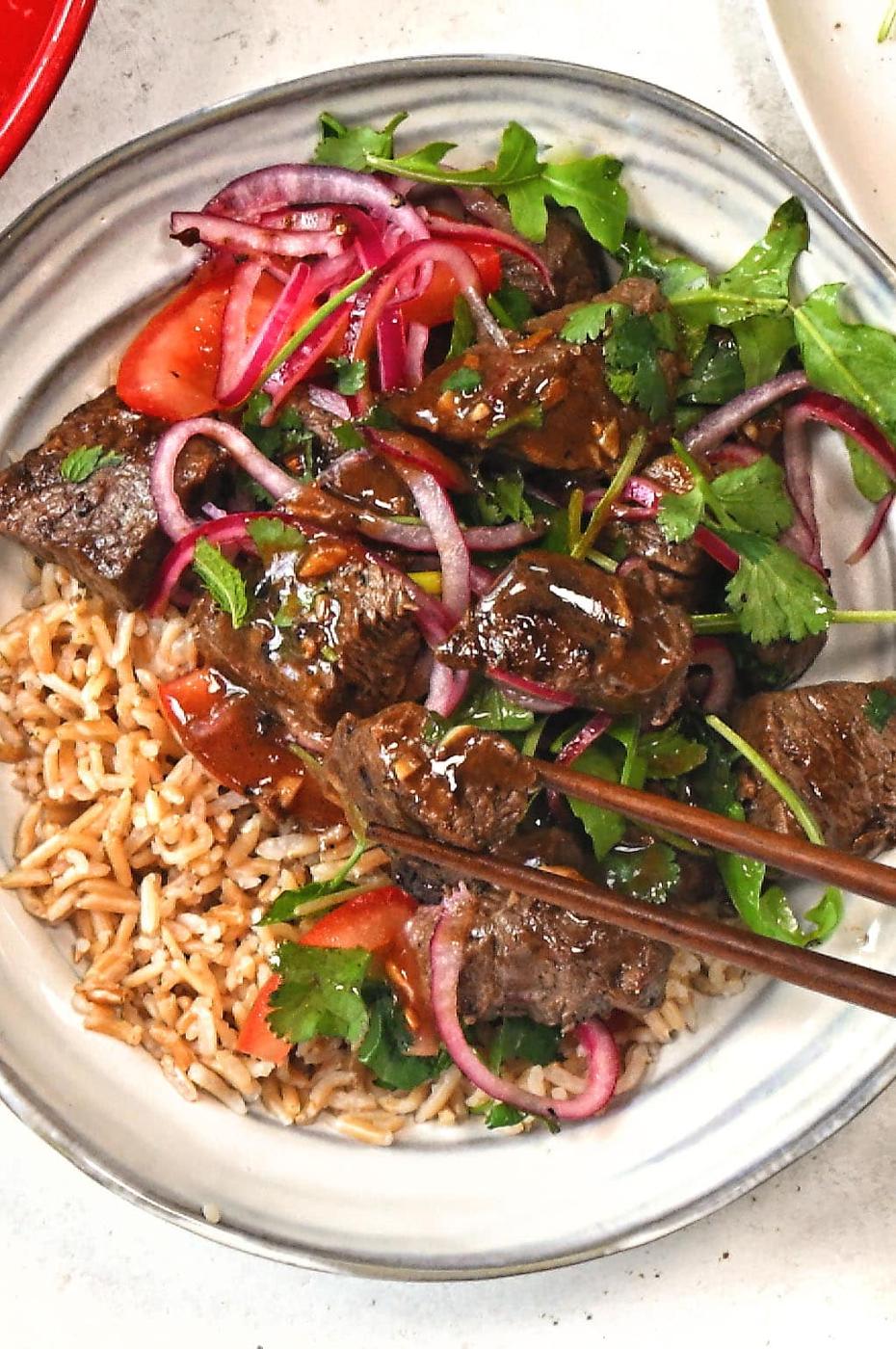 A plate of rice topped with chunks of beef, fresh tomato slices, red onion, and cilantro, with chopsticks picking up a piece of beef. The dish is garnished with a rich brown sauce.