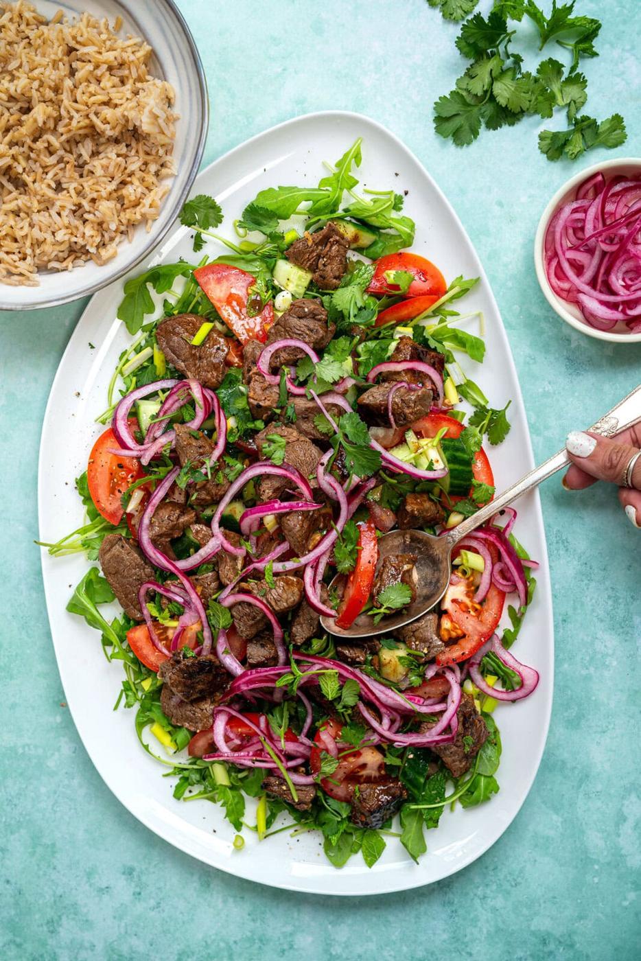 A large white platter with a salad of arugula, tomato wedges, Vietnamese shaking beef, pickled onions, and fresh herbs. A hand holds a spoon, and bowls of brown rice and more red onions sit nearby.