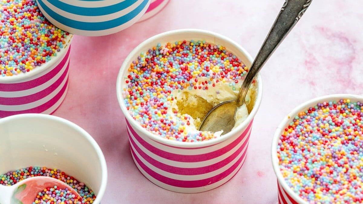 Homemade dot cakes: vanilla cake topped with vanilla buttercream and topped with nonpareil sprinkles, some with a spoon inside. The cups have blue and pink stripes and are arranged on a light pink surface.