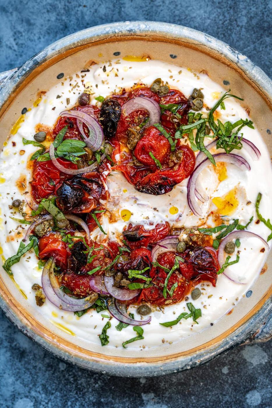 A bowl of creamy cottage cheese dip topped with roasted cherry tomatoes, thinly sliced red onions, capers, and fresh herbs. The dish is drizzled with olive oil and seasoned with pepper flakes, creating a vibrant and colorful presentation.