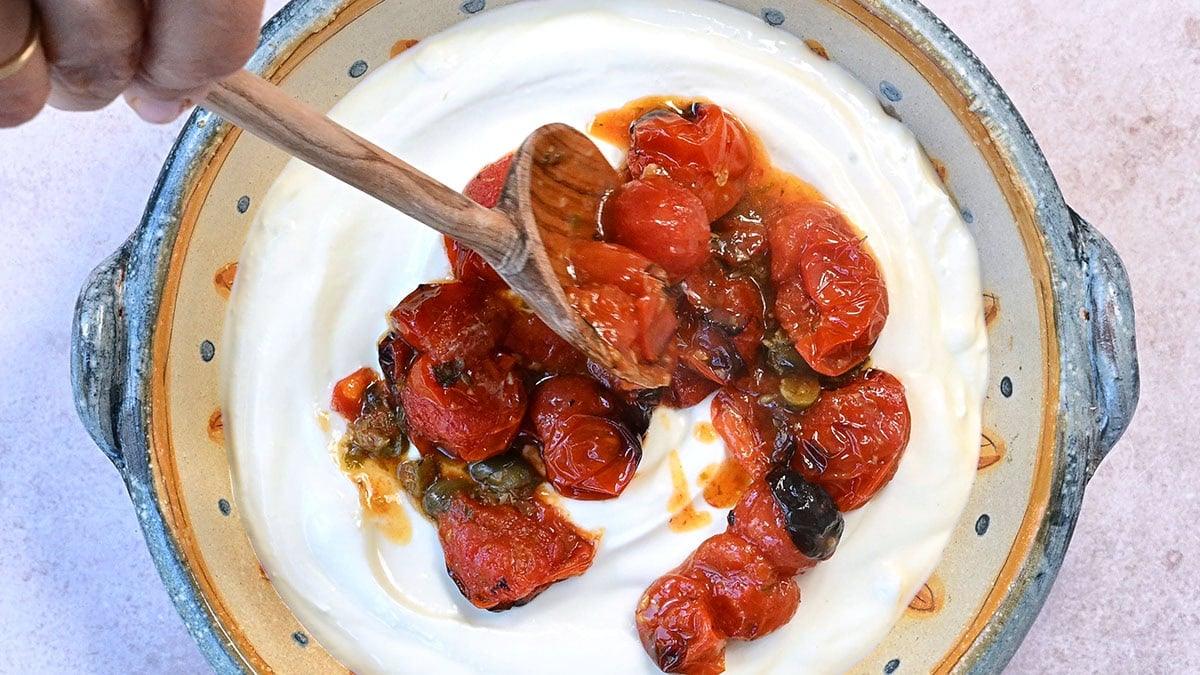 A person using a wooden spoon to spoon roasted cherry tomatoes over whipped cottage cheese in a rustic bowl.