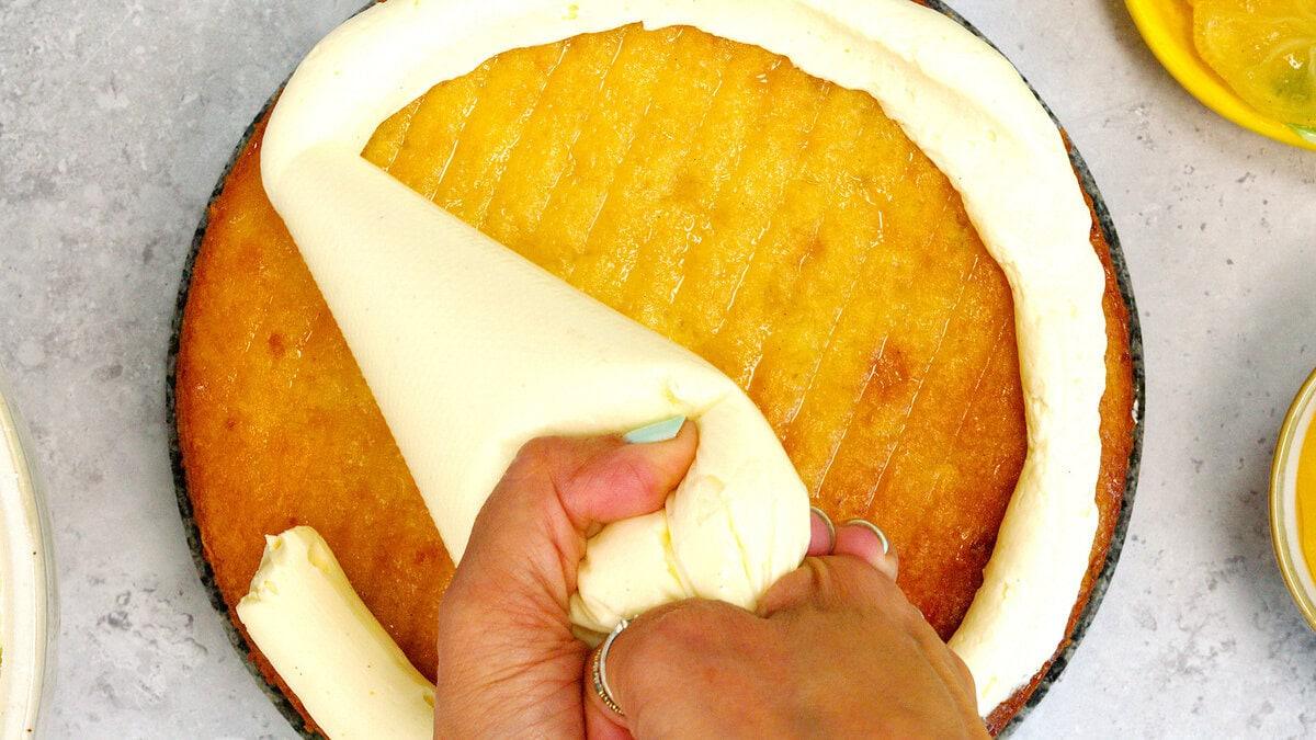 A person pipes a thick ring of white frosting around the edge of a yellow cake layer on a gray surface.