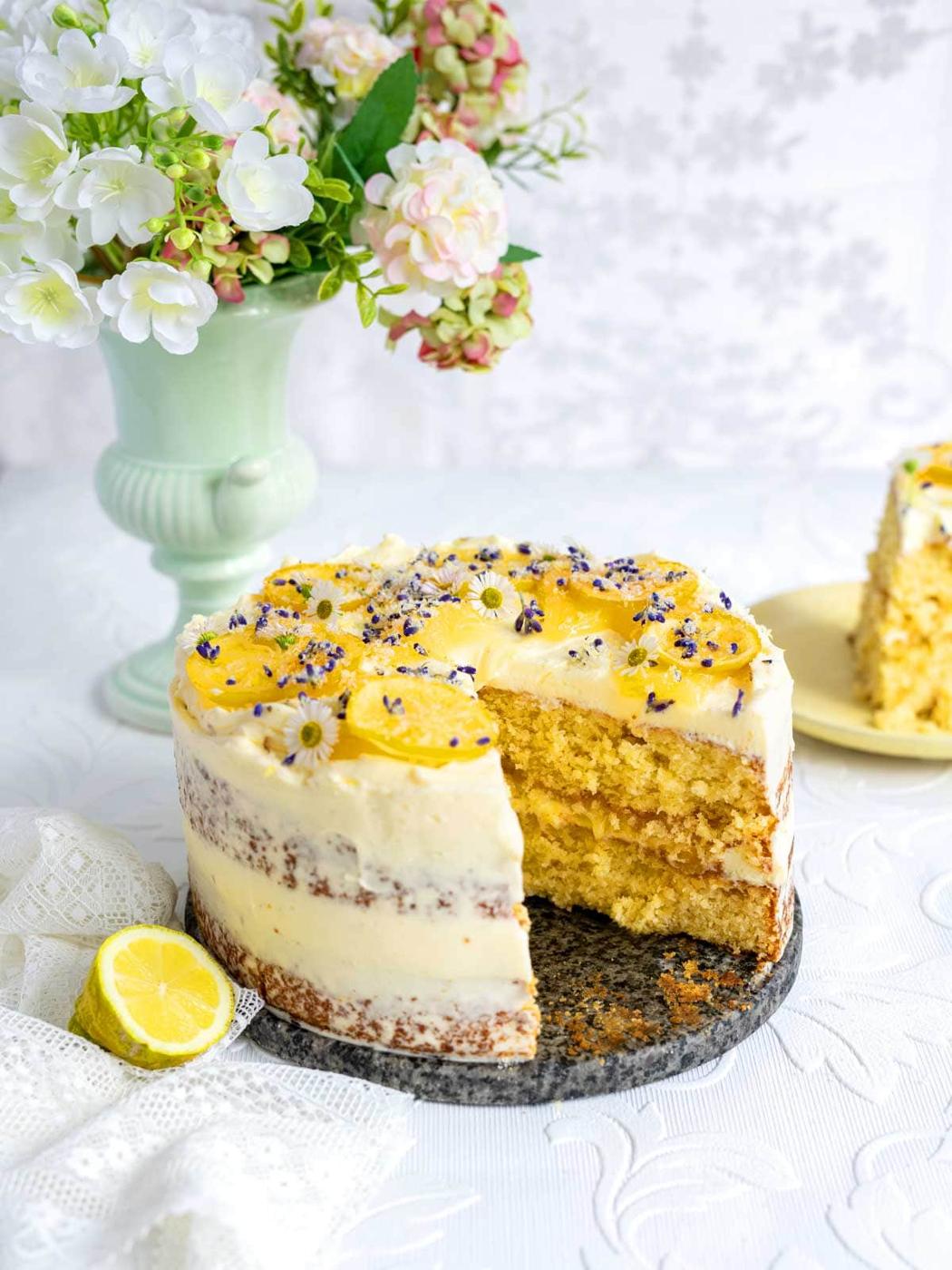 A partially sliced Whole Lemon Cake with creamy frosting and lemon slices on top sits on a marble board. A vase of flowers and a cut lemon are nearby on a white tablecloth.